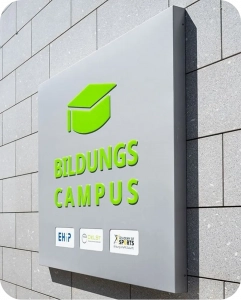 Graues Schild mit grünem Abschlussmützen-Symbol und der Aufschrift ‚Bildungs Campus‘ an einer gefliesten Außenwand; darunter Logos von EHIP, DeLSt und Academy of Sports..