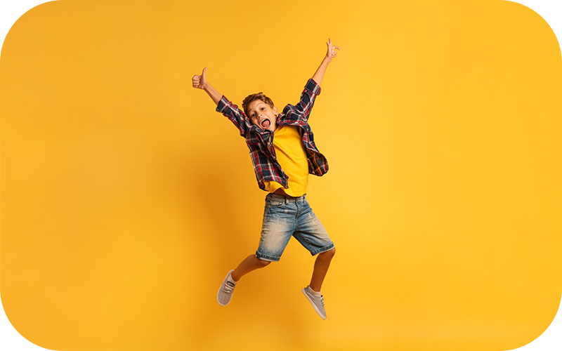 Junge mit kurzen Hosen, kariertem Hemd und gelbem T-Shirt springt vor gelbem Hintergrund mit beiden Armen in der Luft und zeigt Daumen hoch – Symbolbild für fröhliche Kinderaktionen beim Backnanger Kinderfest 2025.