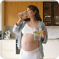 Schwangere Frau steht in heller Küche, trinkt ein Glas Wasser mit Zitrone und hält einen Wasserkrug – gesunde Ernährung und Flüssigkeitszufuhr in der Schwangerschaft.