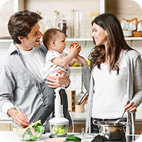 Junge Familie bereitet gemeinsam frisches Essen in der Küche zu. Vater hält Baby, das eine Tomate hält, Mutter steht am Herd – ausgewogene Ernährung für Babys im Familienalltag.
