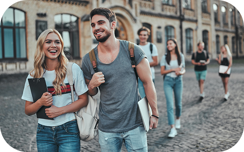 Gruppe junger Studierender läuft lachend über den Campus einer historischen Hochschule. Im Vordergrund ein lächelndes Paar mit Rucksack und Lernmaterialien.
