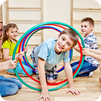 Kinder spielen in einer Turnhalle mit bunten Reifen. Ein Junge krabbelt lachend durch die Reifen, während andere Kinder helfen – Kindersport im Rahmen einer spielerischen Bewegungseinheit.