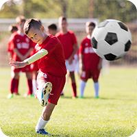 Junge im roten Trikot schießt kraftvoll einen Fußball auf dem Spielfeld, im Hintergrund weitere Kinder in Trainingskleidung – Training im Kinderfußball.
