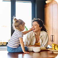 Junge Familie bereitet gemeinsam frisches Essen in der Küche zu. Vater hält Baby, das eine Tomate hält, Mutter steht am Herd – ausgewogene Ernährung für Babys im Familienalltag durch vegane Ernährungsberatung.