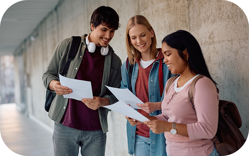 Drei junge Studierende stehen in einem modernen Hochschulgebäude und schauen gemeinsam lächelnd auf Unterlagen – symbolisiert Austausch und gemeinsames Lernen.