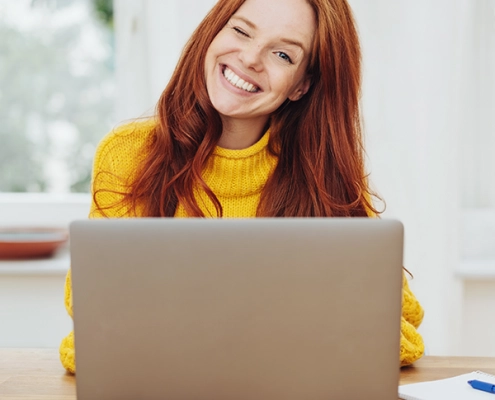 Junge Frau nimmt lächelnd an einer Online-Infoveranstaltung teil, sitzt mit Laptop in modernem, hellen Raum.