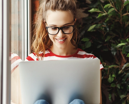 Junge Frau nimmt lächelnd an einer Online-Infoveranstaltung teil, sitzt mit Laptop in modernem, hellen Raum.