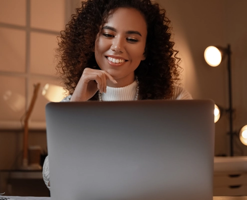 Junge Frau nimmt lächelnd an einer Online-Infoveranstaltung teil, sitzt mit Laptop in modernem, hellen Raum.