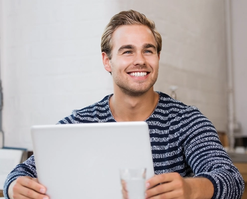 Junger Mann nimmt lächelnd an einer Online-Infoveranstaltung teil, sitzt mit Laptop in modernem, hellen Raum.