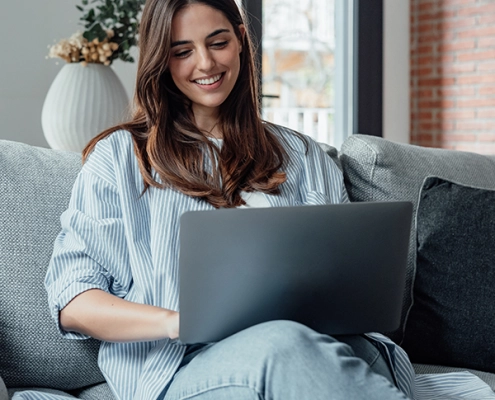 Junge Frau nimmt lächelnd an einer Online-Infoveranstaltung teil, sitzt mit Laptop in modernem, hellen Raum.